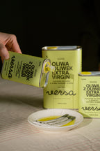 A hand pours extra virgin olive oil from a green tin labeled Oliwa z oliwek extra virgin versa into a white dish with two spoons, with two similar tins in the background.