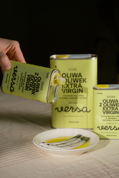 A hand pours extra virgin olive oil from a green tin labeled Oliwa z oliwek extra virgin versa into a white dish with two spoons, with two similar tins in the background.