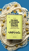 Puszka oliwy z oliwek extra virgin versa o jasnozielonej etykiecie leży na kremowej siatce wśród zielonych oliwek, na intensywnie niebieskim tle. Na etykiecie widoczny napis: „Oliwa z oliwek extra virgin”, „Tłoczona na zimno z jednej odmiany oliwek” oraz logo versa.